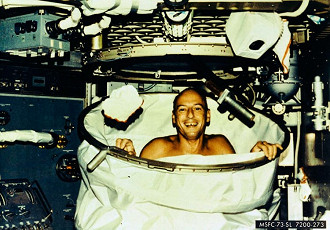 Charles Conrad, Jr., comandante do Skylab-2, sorri depois de um banho quente no chuveiro nos aposentos da tripulação do Orbital Workshop da estação espacial Skylab. Ao implantar o chuveiro, a cortina do chuveiro foi puxada do chão e presa ao teto. A água veio através de um chuveiro com botão de pressão ligado a uma mangueira flexível. A água foi retirada por um sistema de vácuo.